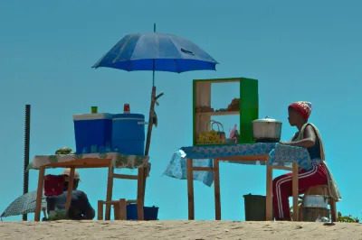 Two vendors sit behind colorful roadside food and drink stalls on a sandy hilltop in Madagascar, set against a clear turquoise sky.
